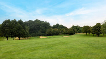 Green golf course with sand bunkers, trees, and a blue sky with light clouds.