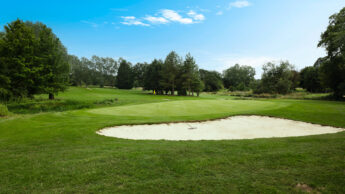 A golf course with a sand bunker and green, surrounded by trees under a blue sky.