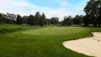 Golf green with flagstick, surrounded by trees and a sand bunker on the right side.