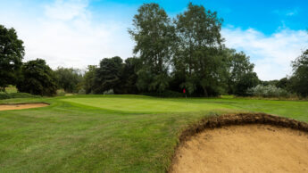 Golf course green with sand bunker in foreground and trees in the background under a blue sky.