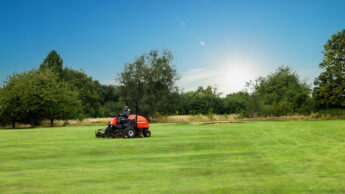Person driving a red lawnmower on a large grassy field under a clear blue sky.
