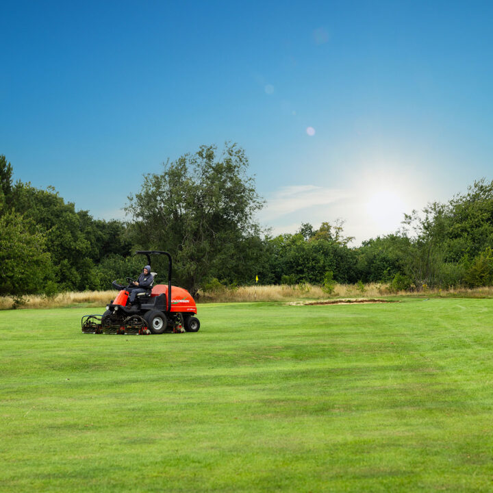 Person driving a red lawnmower on a large grassy field under a clear blue sky.