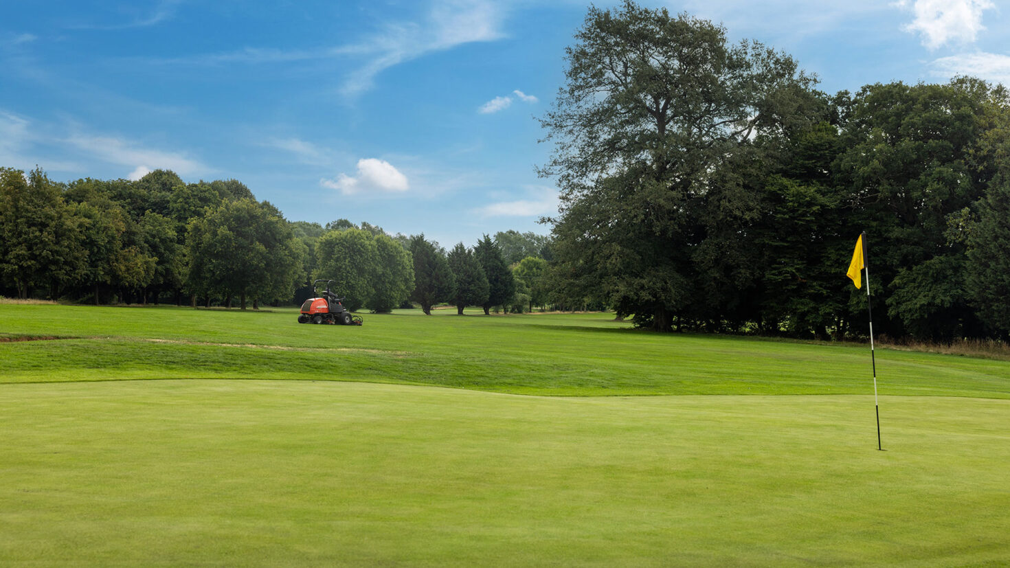 A golf course with a yellow flag on the green and a mower in the distance under a blue sky.