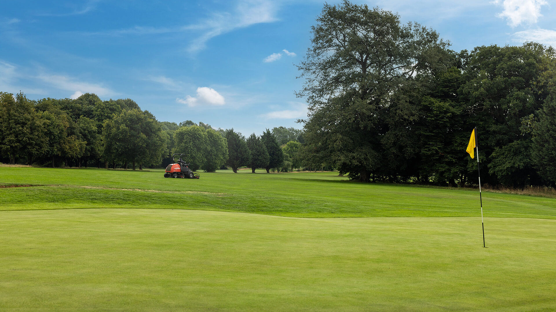 A golf course with a yellow flag on the green and a mower in the distance under a blue sky.