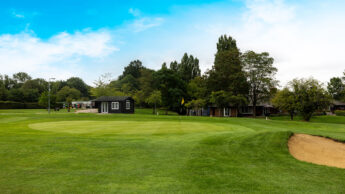 Golf green with a flag, sand bunker, and small buildings surrounded by trees under blue sky.