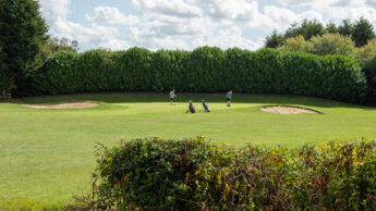 Three people golfing on a green with sand bunkers, trees, and bushes in the background.