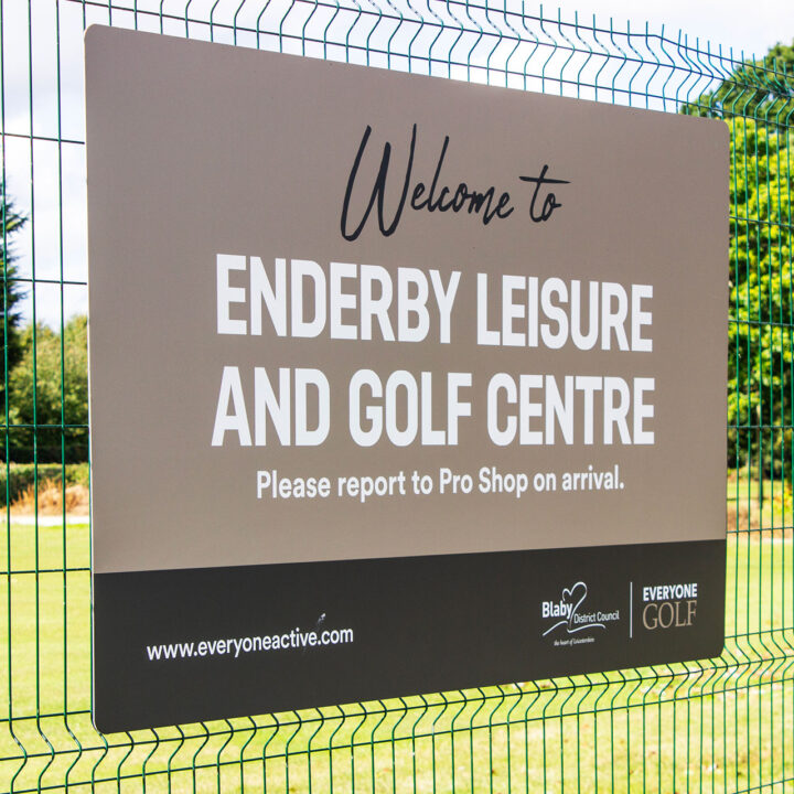 Sign reading "Welcome to Enderby Leisure and Golf Centre" on a fence by a green field.