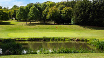A golf green with a flag, bordered by trees and a pond in the foreground.