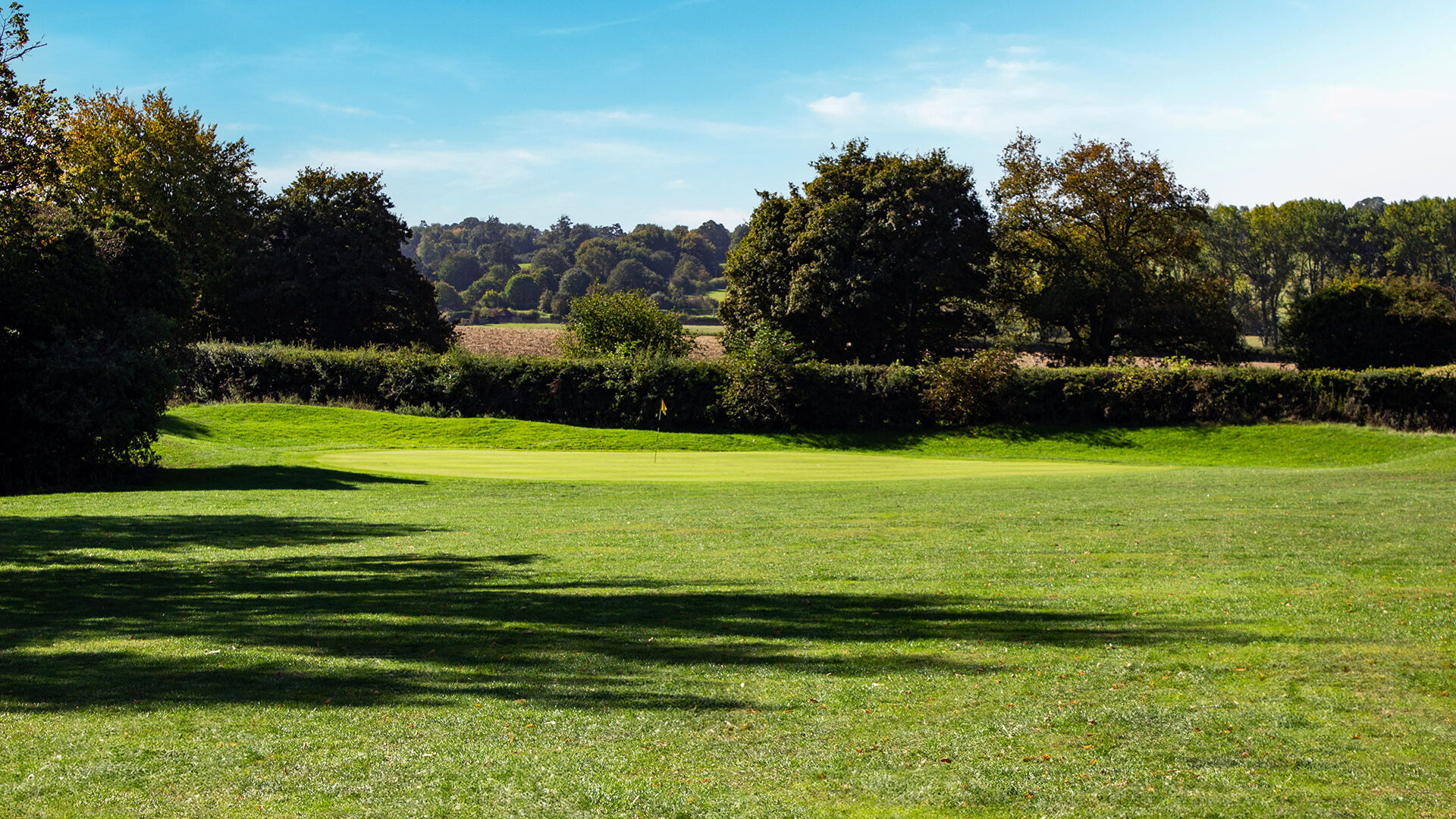 A green golf course with trees and hills under a clear blue sky.