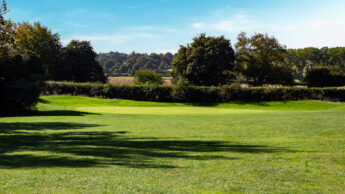 A green golf course with trees and hills under a clear blue sky.