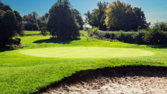 A golf green with a flagstick, bordered by trees and a sand bunker under a clear sky.
