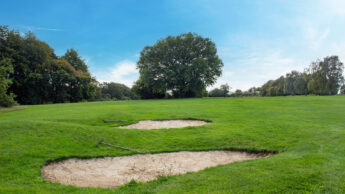 Two sand bunkers on a green golf course under a blue sky, surrounded by trees.