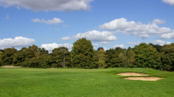 A green golf course with sand bunkers and trees under a blue sky with clouds.