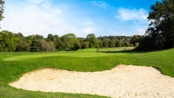 A golf course with a sand bunker, green grass, and trees under a blue sky.
