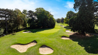 A golf green surrounded by sand bunkers and trees under a bright blue sky.