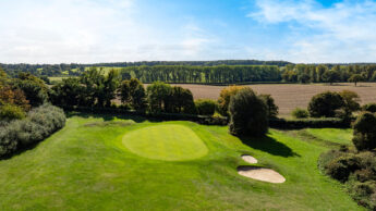 A golf green with a sand bunker, surrounded by trees and open countryside under a blue sky.