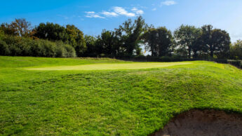 Golf course with green grass, a sand bunker, and trees under a clear blue sky.
