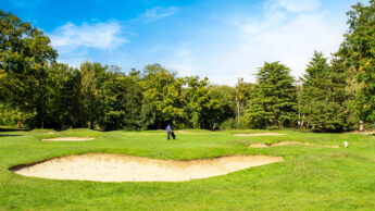 A golfer walks on a green golf course near sand bunkers under a blue sky.