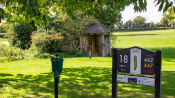 Golf course hole 18 sign, green grass, trees, and a small wooden shelter in the background.