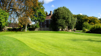 Sunny golf course with a flag on the green, trees, and a brick house in the background.