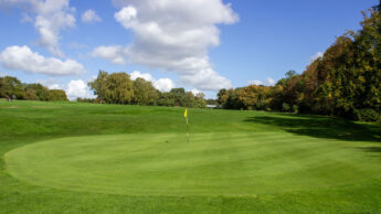 A golf green with a yellow flag under a blue sky with scattered clouds.