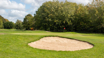A sand bunker on a golf course with a green and flag in the background, surrounded by trees.