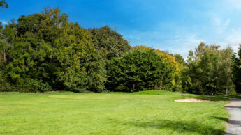 A golf course with green grass, a sand bunker, trees, and a clear blue sky.
