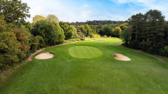 A green golf course with two sand bunkers and trees in the background under a blue sky.