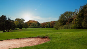 A golf course with a sand bunker, green grass, trees, and a bright sky at sunset.