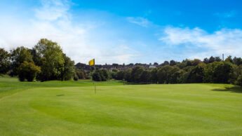 A sunny golf course with a yellow flag on the green and trees in the background.
